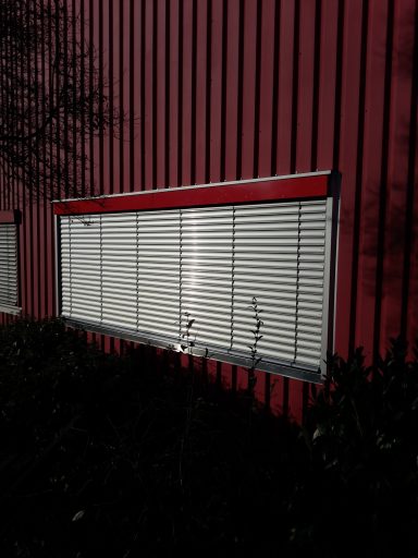 Rote Wand mit geschlossenen Jalousien und Pflanzen davor.