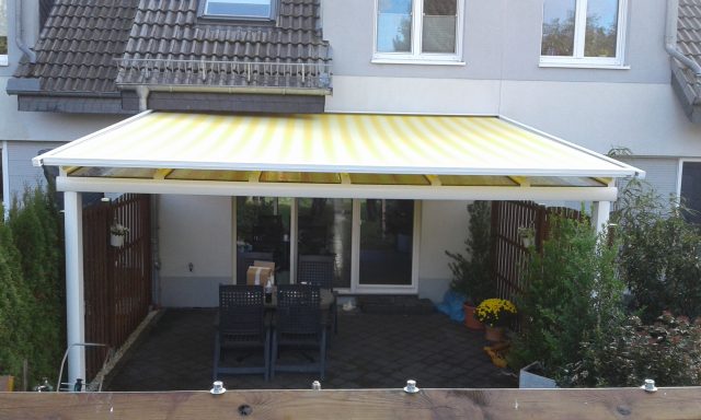 Gelbe Markise über einer Terrasse mit Sitzgruppe und grünen Pflanzen im Hintergrund. Überdachung mit Obermarkise